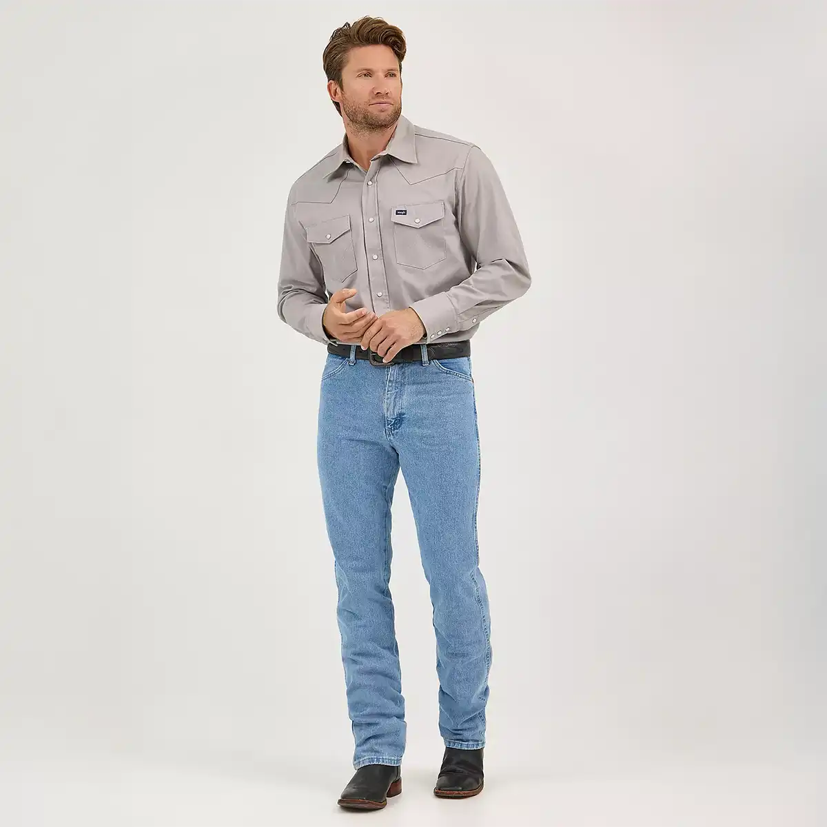 Full length front view of a man wearing Wrangler Men's Cowboy Cut 0936 slim fit antique wash jeans with grey Western shirt and black boots.
