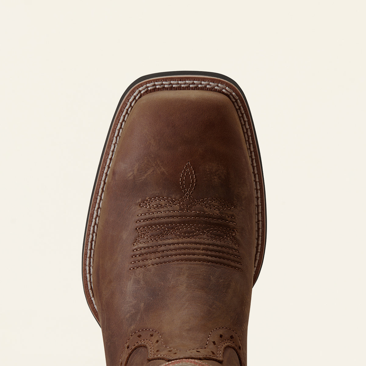 A close-up top-down view of a brown leather cowboy boot toe, showing the square vamp, decorative stitching, and embossed floral pattern against a light cream background.