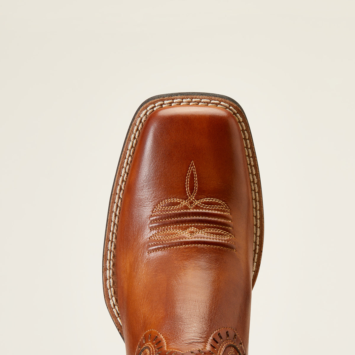Close-up of the square toe of a brown leather Ariat western boot with decorative stitching and a square toe, shown against a light beige background.