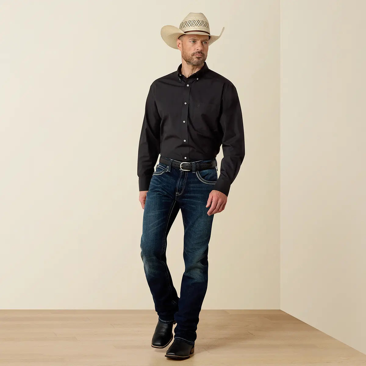 Full-length lifestyle view of the model wearing the Ariat black wrinkle-free shirt with blue jeans, black boots, and a straw cowboy hat.