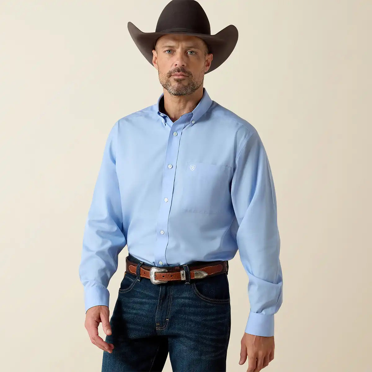 Man wearing a light blue Ariat shirt, dark jeans, and a cowboy hat against a beige background.