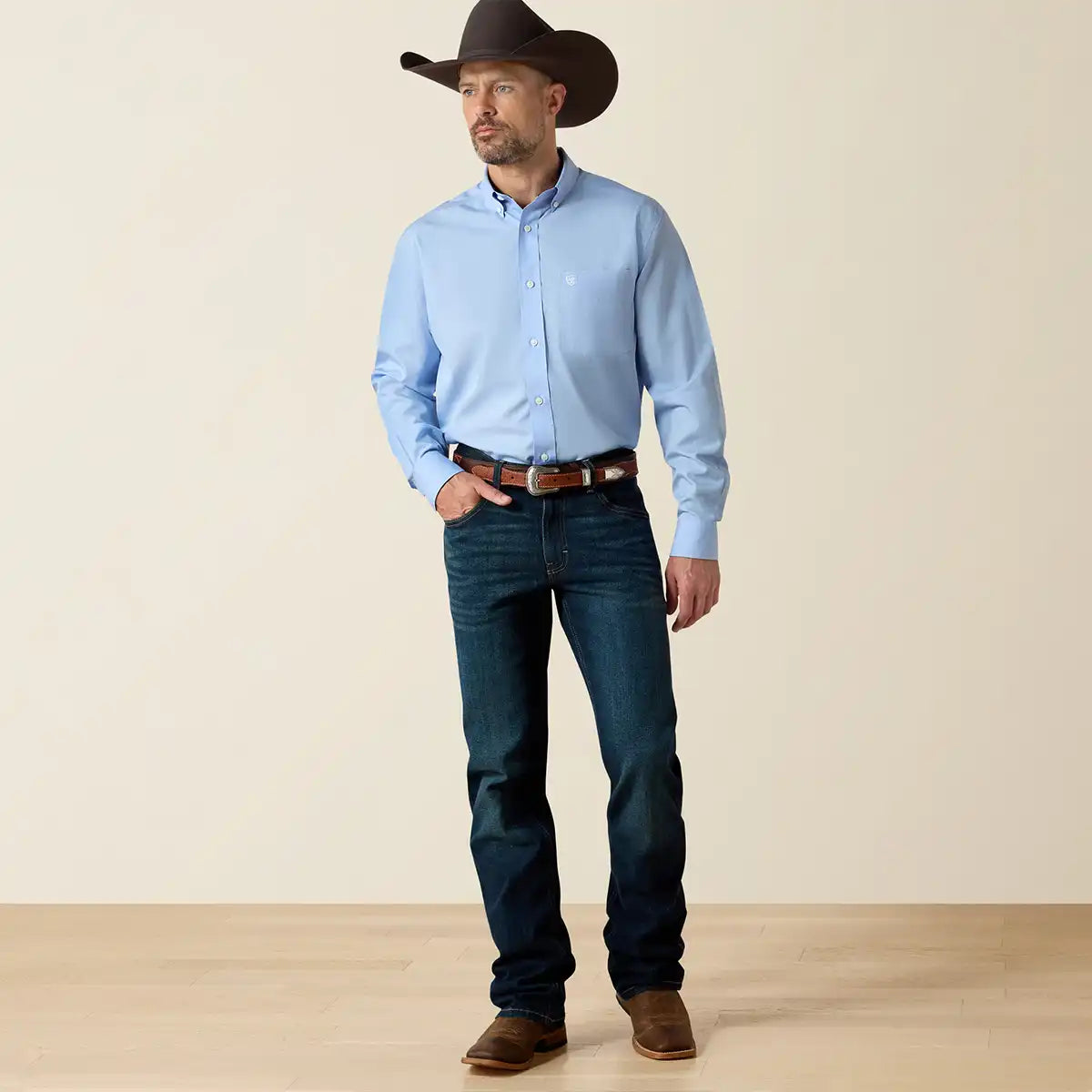 Man wearing a blue Ariat shirt, dark jeans, and a cowboy hat against a beige background.