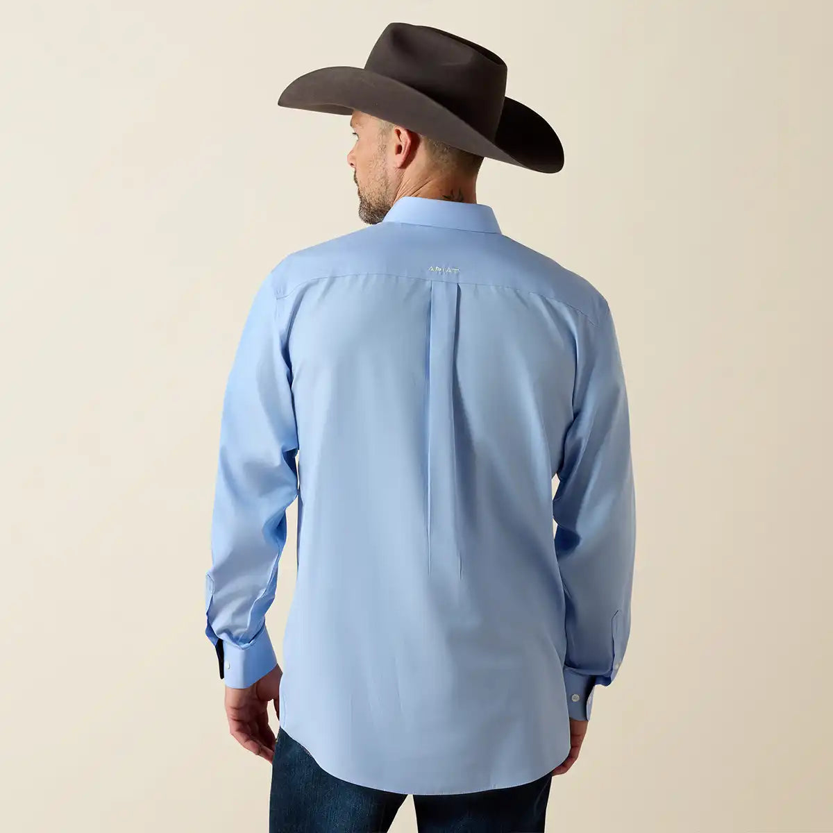 A man wearing a light blue long-sleeve button-down Ariat shirt and a dark brown cowboy hat, shown from the back against a plain beige studio background.