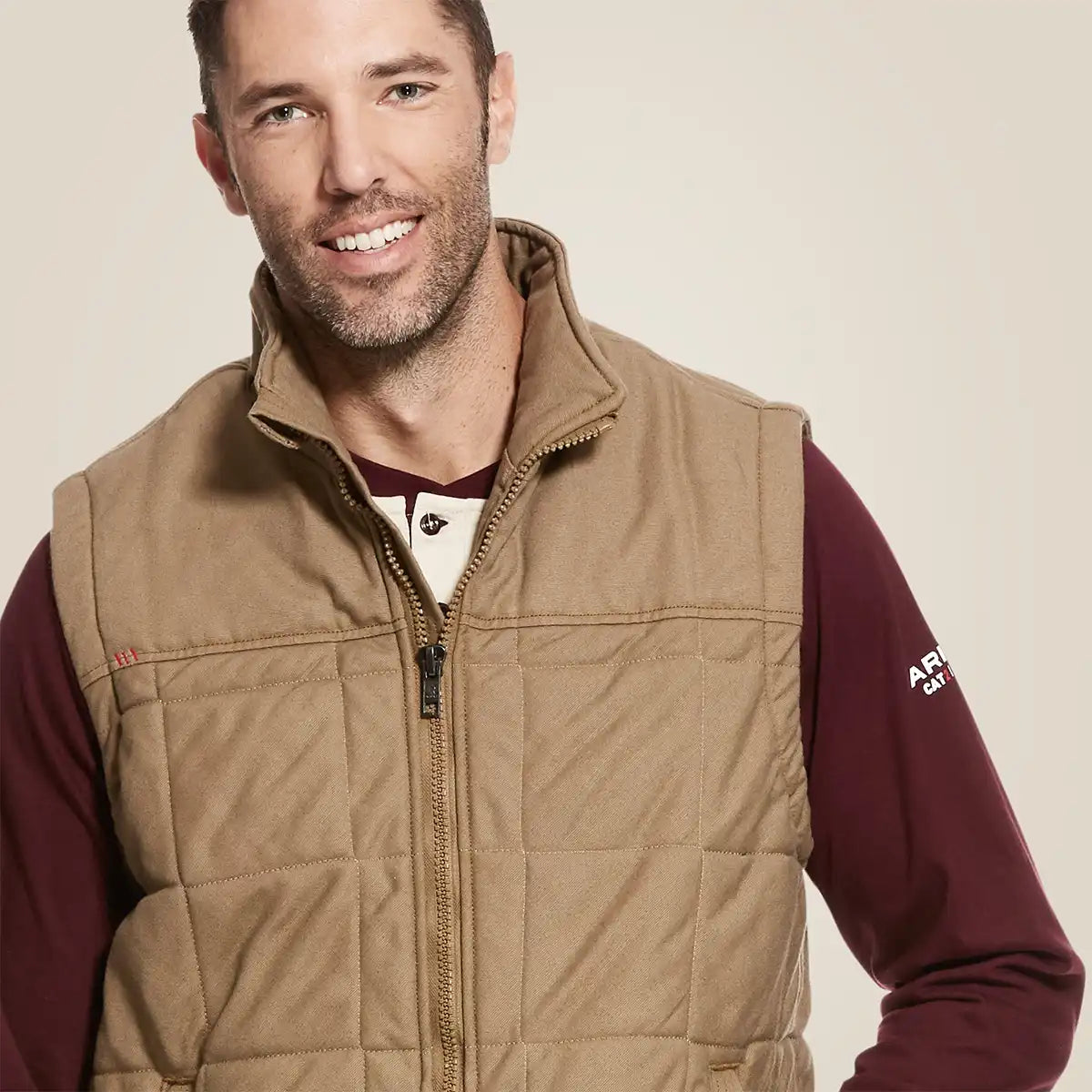 A smiling man wearing a tan quilted zip-front Ariat vest over a burgundy long-sleeve shirt, photographed against a plain light beige studio background.