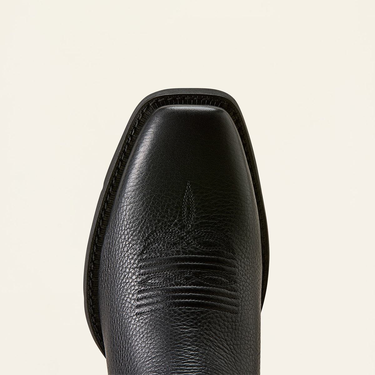 Close-up of the square toe of a black leather Ariat cowboy boot, showing textured leather and decorative stitching against a light cream background.