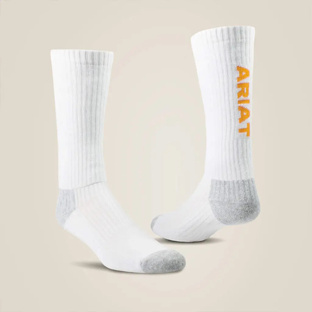A pair of white crew-length boot socks displayed against a light beige background, featuring grey heel and toe panels, ribbed uppers, and orange vertical "ARIAT" text printed along the side of the leg.