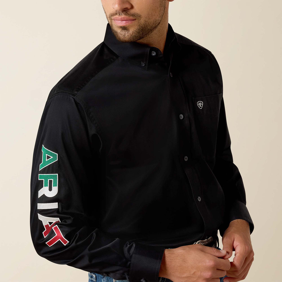 A man wearing a black long-sleeve button-up shirt with a large vertical 'ARIAT' graphic in green, white, and red on the right sleeve, and a small shield logo on the left chest pocket, against a plain beige background.