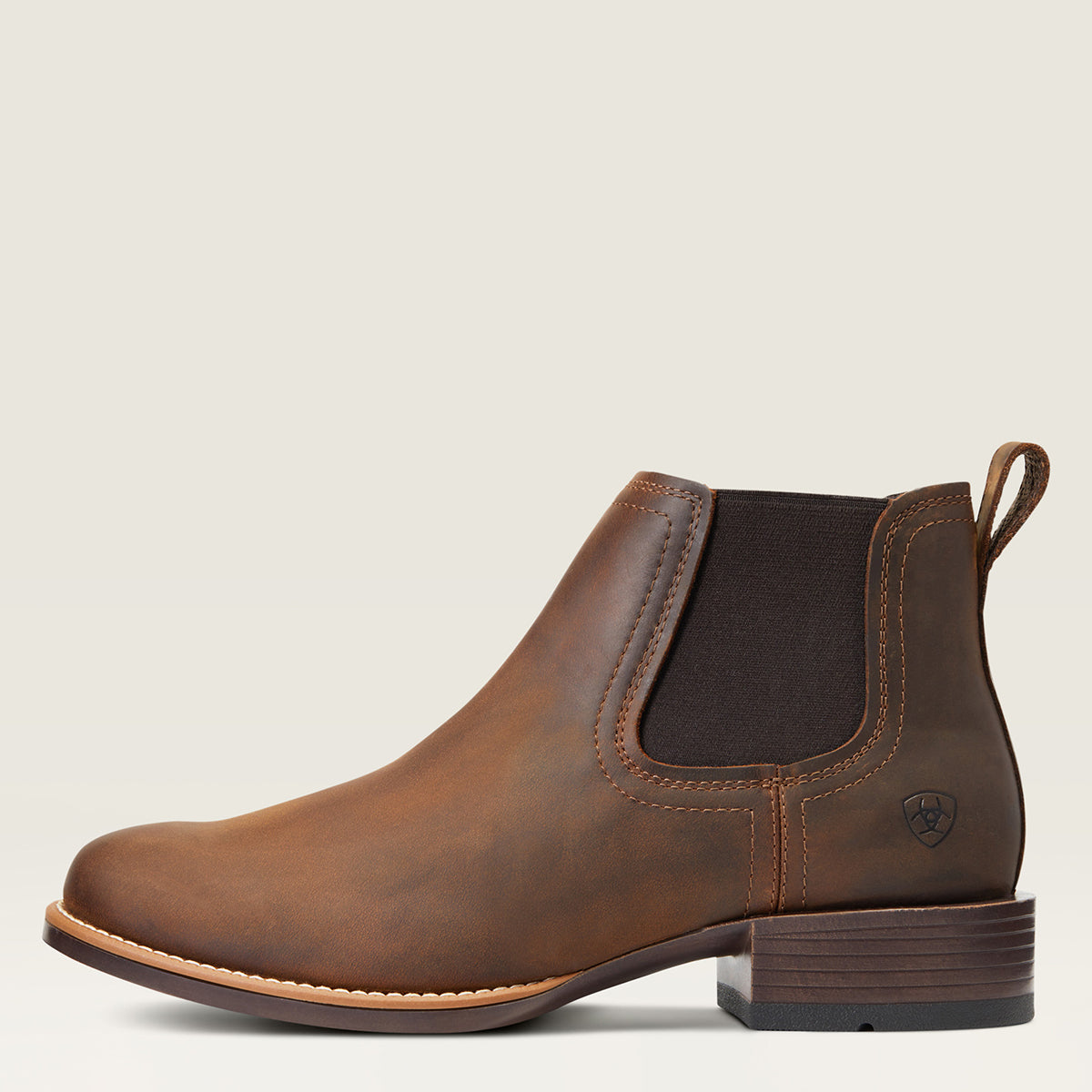 A brown leather Ariat Chelsea ankle boot with a dark brown elastic side panel and a low stacked heel, shown in profile against a light beige background.