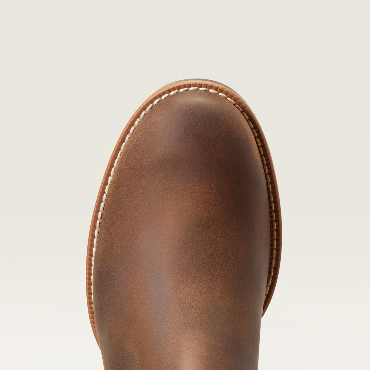 Close-up of the Ariat round toe of a brown leather boot with white contrast stitching, shown against a light off-white background.