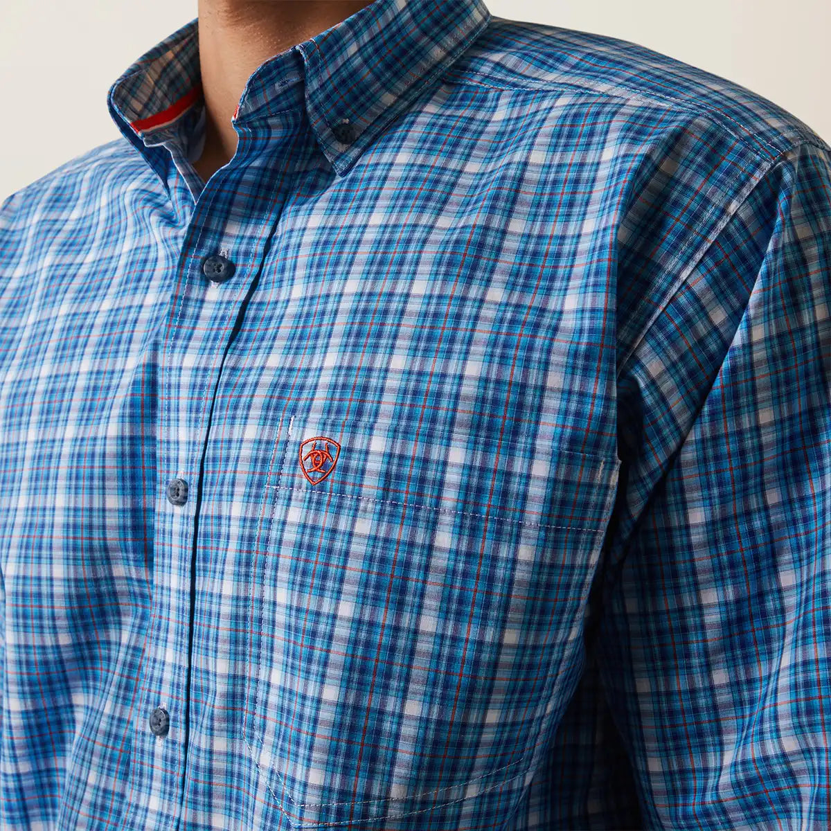 A close-up of a men's blue and white plaid button-down shirt with a small red embroidered shield Ariat logo on the chest pocket. The shirt is worn by a model, with the neck and collar visible.