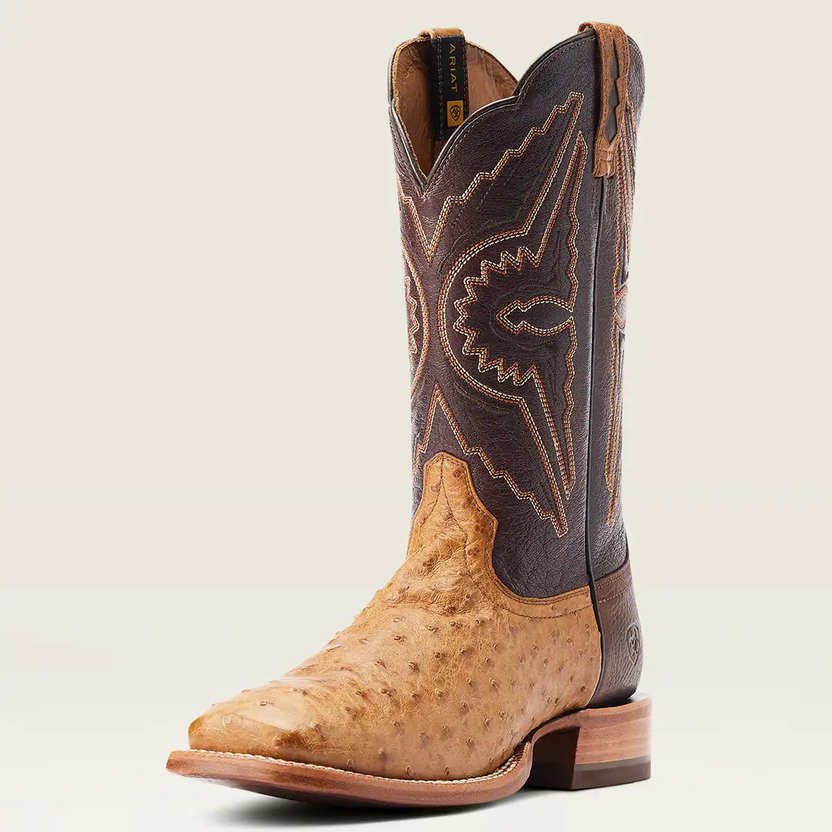 A single Ariat western cowboy boot shown in a side profile on a light beige background. The boot features a tan ostrich leather foot, a dark brown leather shaft with tan embroidered stitching, and a stacked wood-look heel.