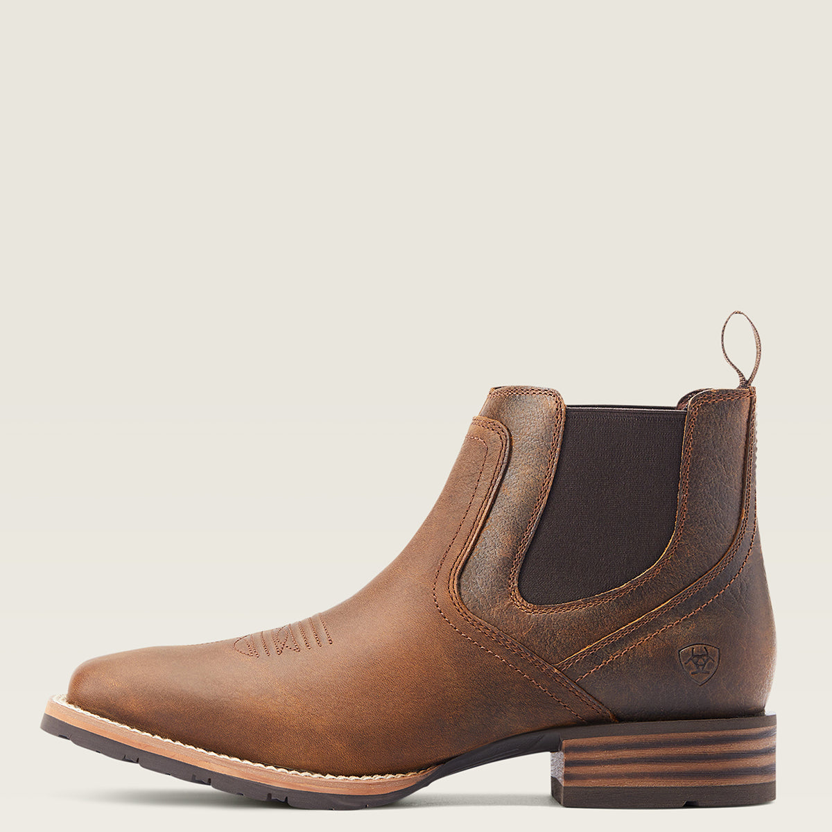 Ariat men's brown leather Chelsea-style ankle boot with elastic side gore panels, a pull loop, and a stacked block heel, shown in side profile on a light warm gray background.