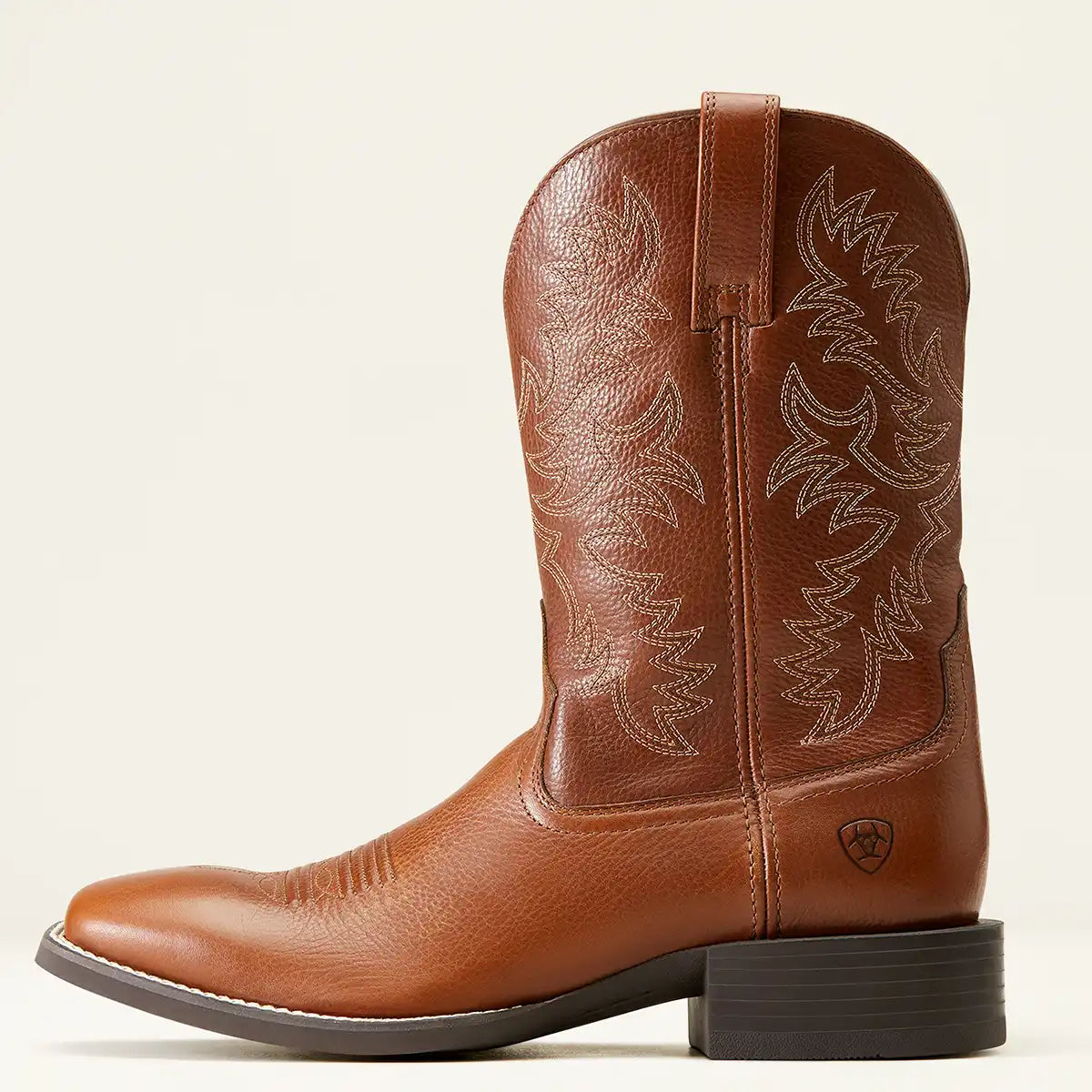 A single brown leather Ariat cowboy boot with western embroidery on the shaft, shown in profile against a light beige background. The boot features a dark brown block heel and a small shield logo near the heel.