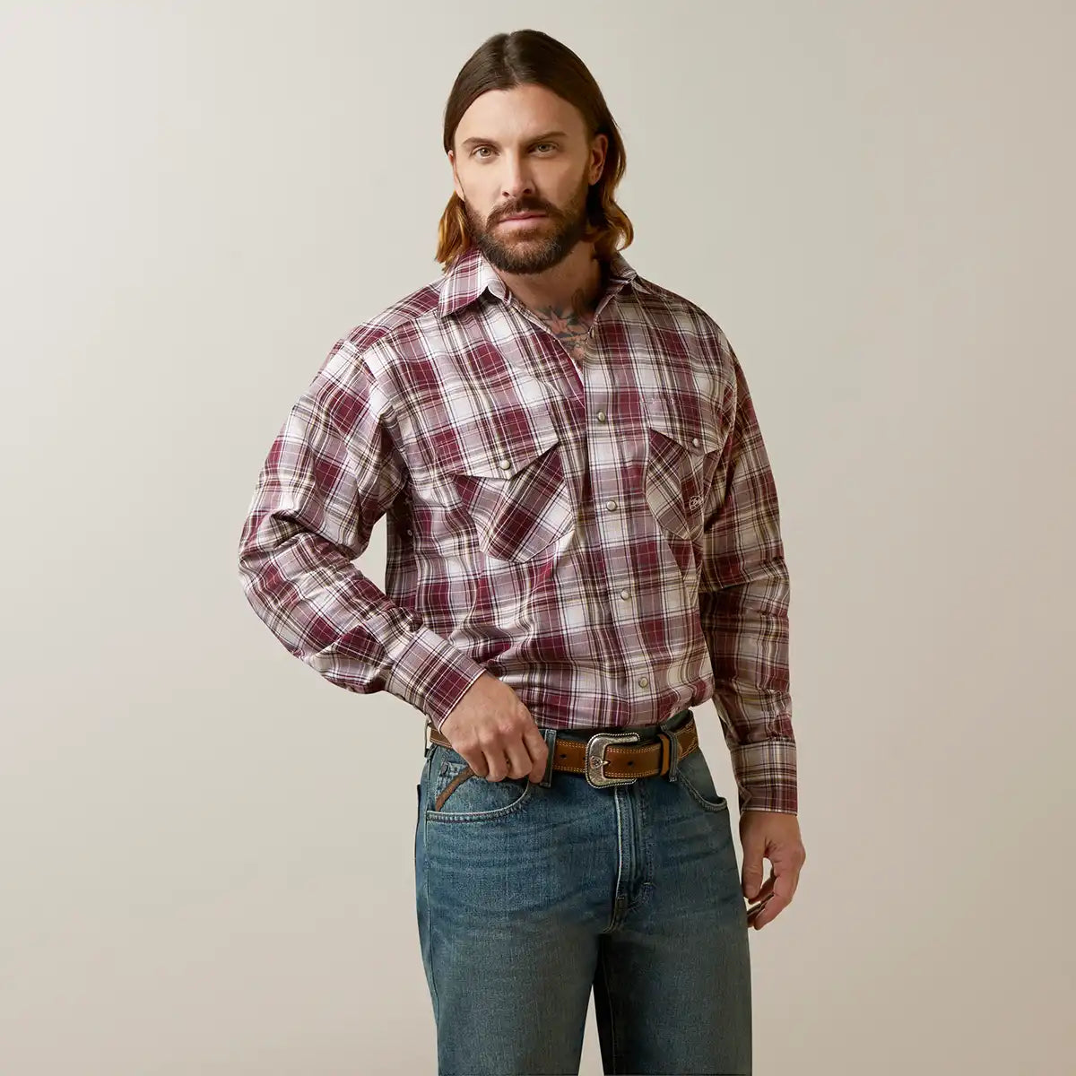 A man wearing a long-sleeve western-style plaid button-down Ariat shirt in burgundy and beige, paired with blue jeans and a brown belt, standing against a plain light gray background.