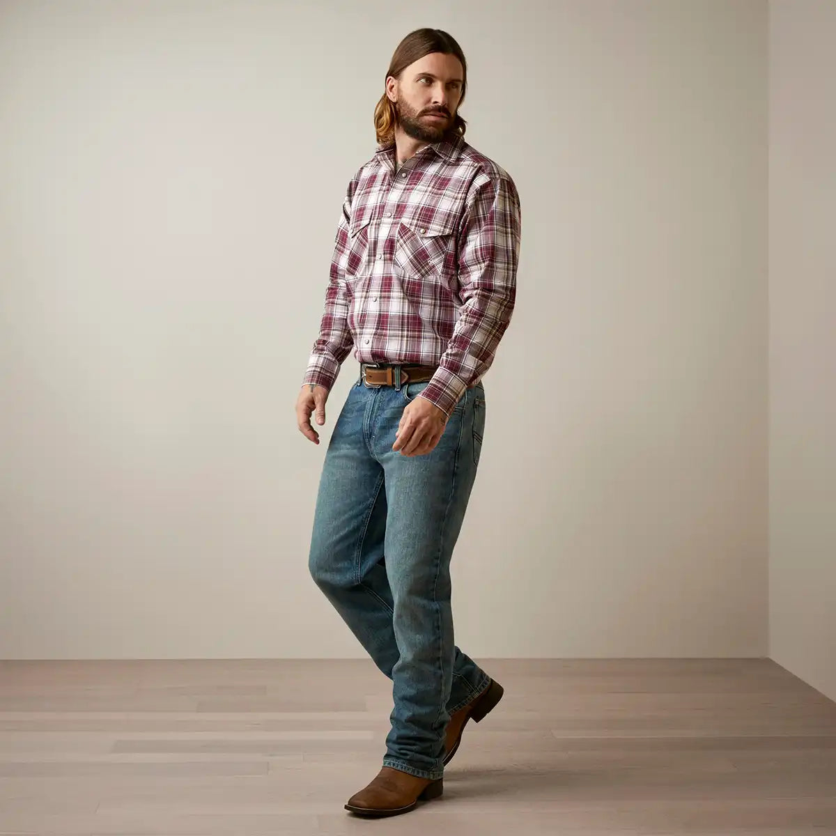 A man wearing a long-sleeve plaid button-down Ariat shirt in red and white, blue jeans, a brown belt, and brown boots, standing in a minimalist studio with a beige wall and light wood floor.