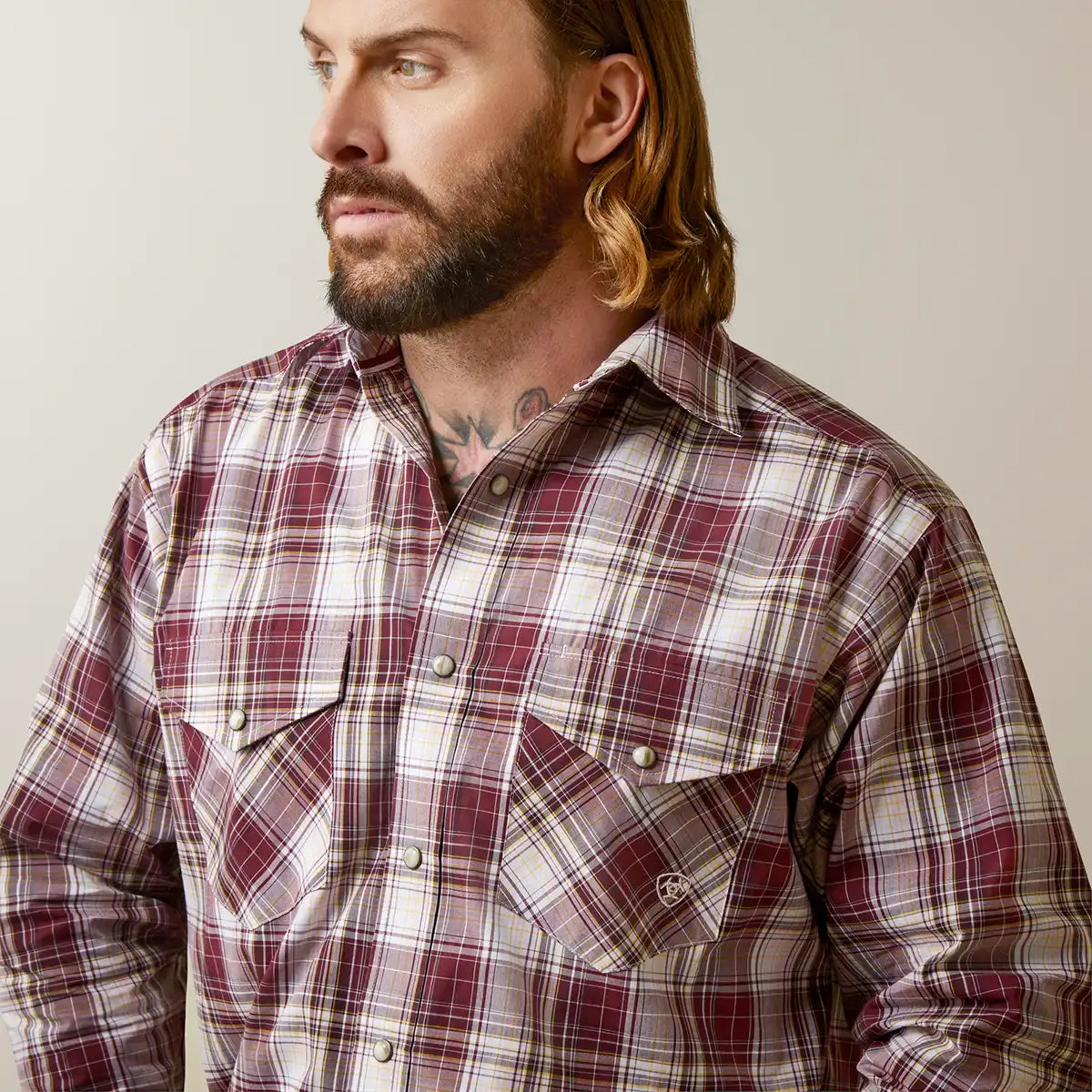 A man wearing a long-sleeve button-up plaid shirt in burgundy, beige, and white, photographed against a plain light gray studio background.