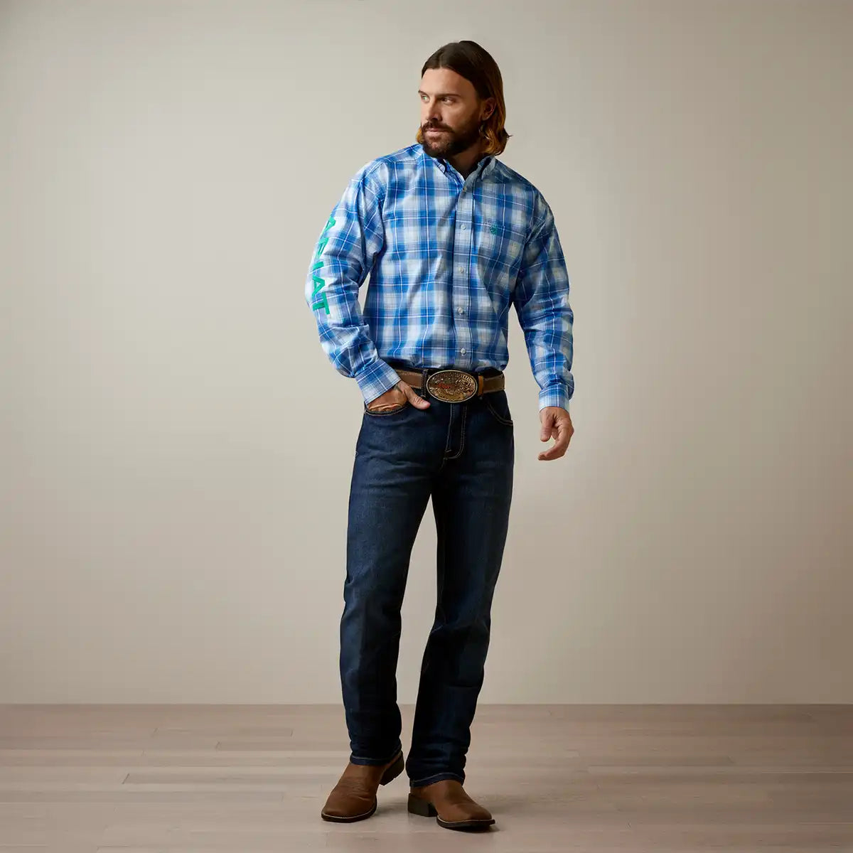 A man wearing a blue and white plaid long-sleeve button-down Ariat shirt, dark blue jeans, a brown belt with a large decorative buckle, and brown boots, standing against a plain beige studio background.