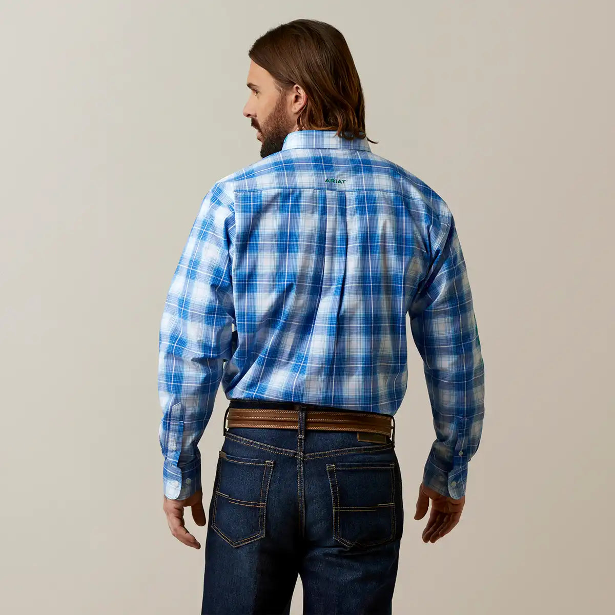A man viewed from the back wearing a blue and white plaid long-sleeve Ariat shirt, dark blue jeans, and a brown belt, against a plain light beige studio background.