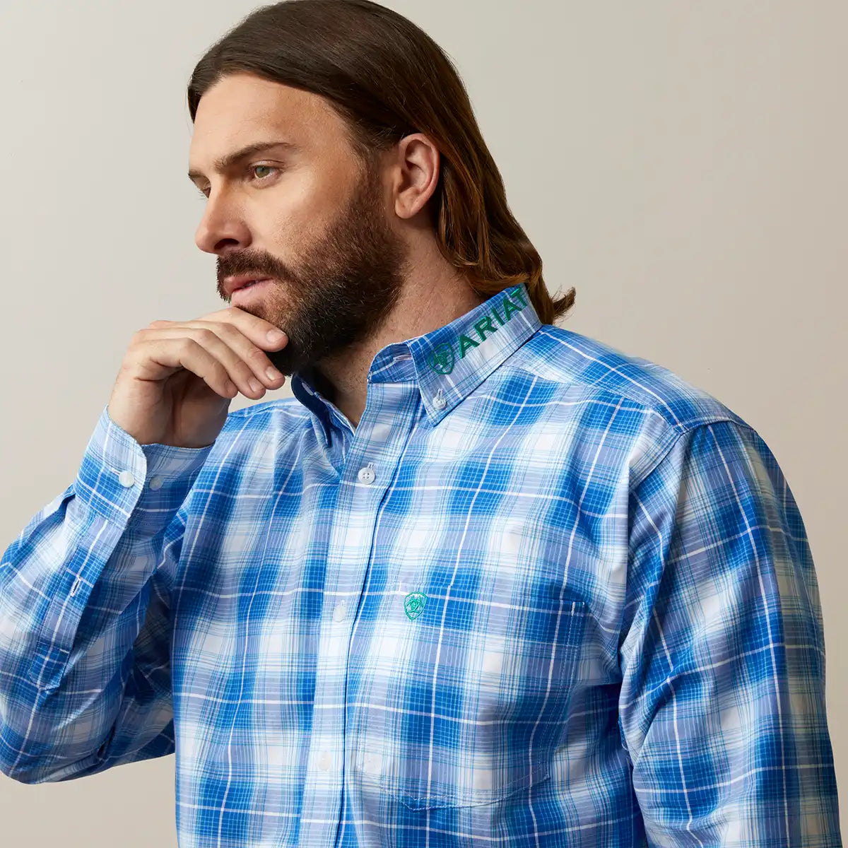 A man wearing a blue and white plaid button-down shirt with green embroidered details on the collar and chest, posed against a neutral beige background.