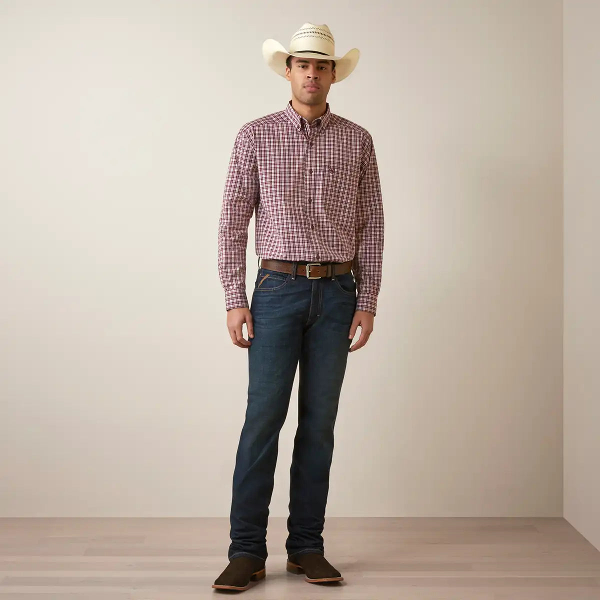 A man wearing a red and white gingham long-sleeve button-down Ariat shirt, dark blue jeans, a brown belt, brown boots, and a cream cowboy hat, standing against a plain beige wall.