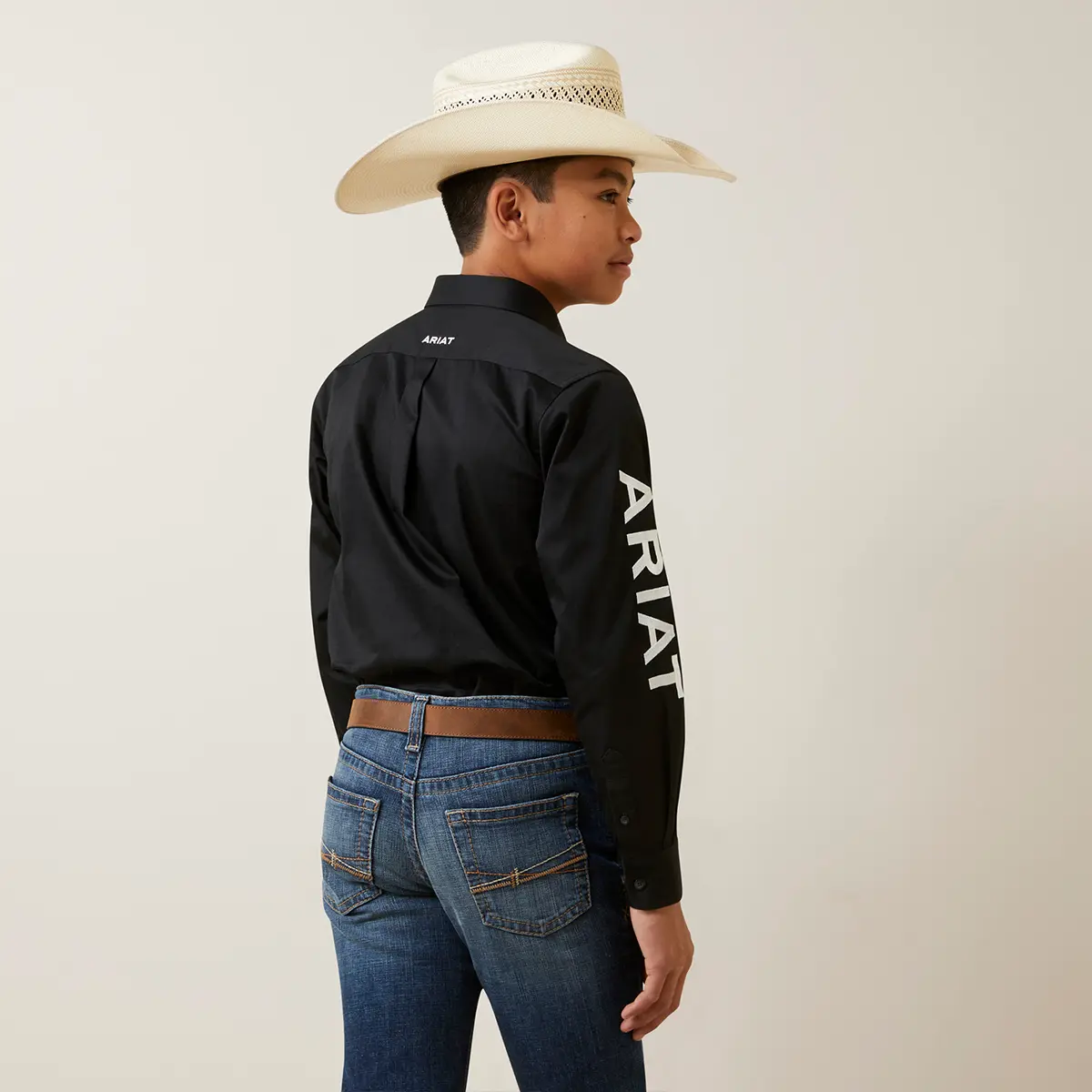 Rear three-quarter studio view of the boy showcasing the bold white ARIAT text embroidered on the right sleeve and back yoke logo.