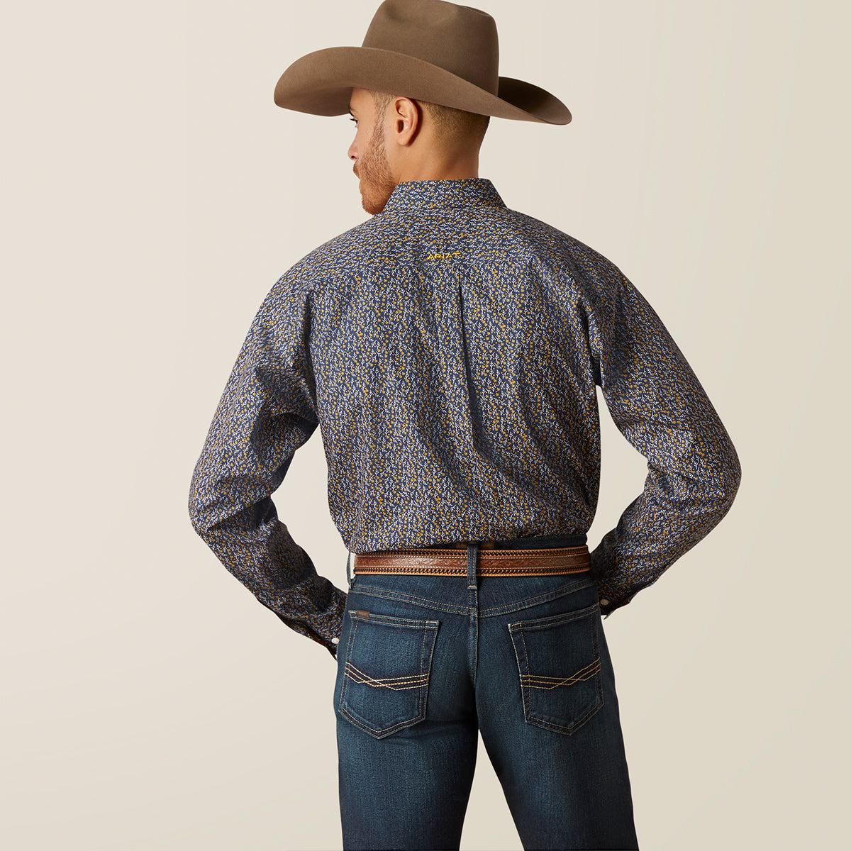 A man wearing a brown cowboy hat, a long-sleeve Ariat western shirt with a blue, yellow, and orange flowers print, and blue jeans, shown from the back against a light beige background.