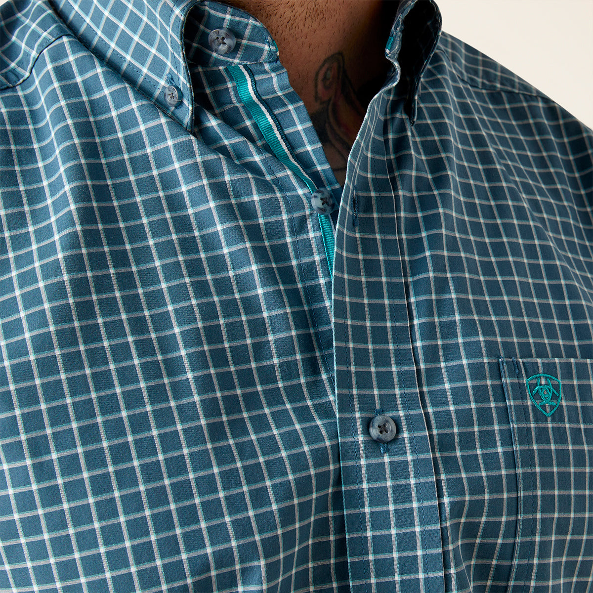 Close-up of a men's button-down shirt with a blue and white windowpane check pattern, featuring a small embroidered logo on the chest pocket.