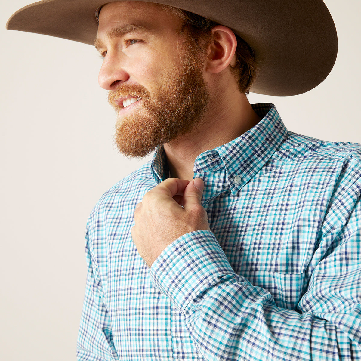 A man wearing a brown cowboy hat and a blue, white, and black checkered long-sleeve button-down shirt, adjusting the collar of his shirt against a plain light background.