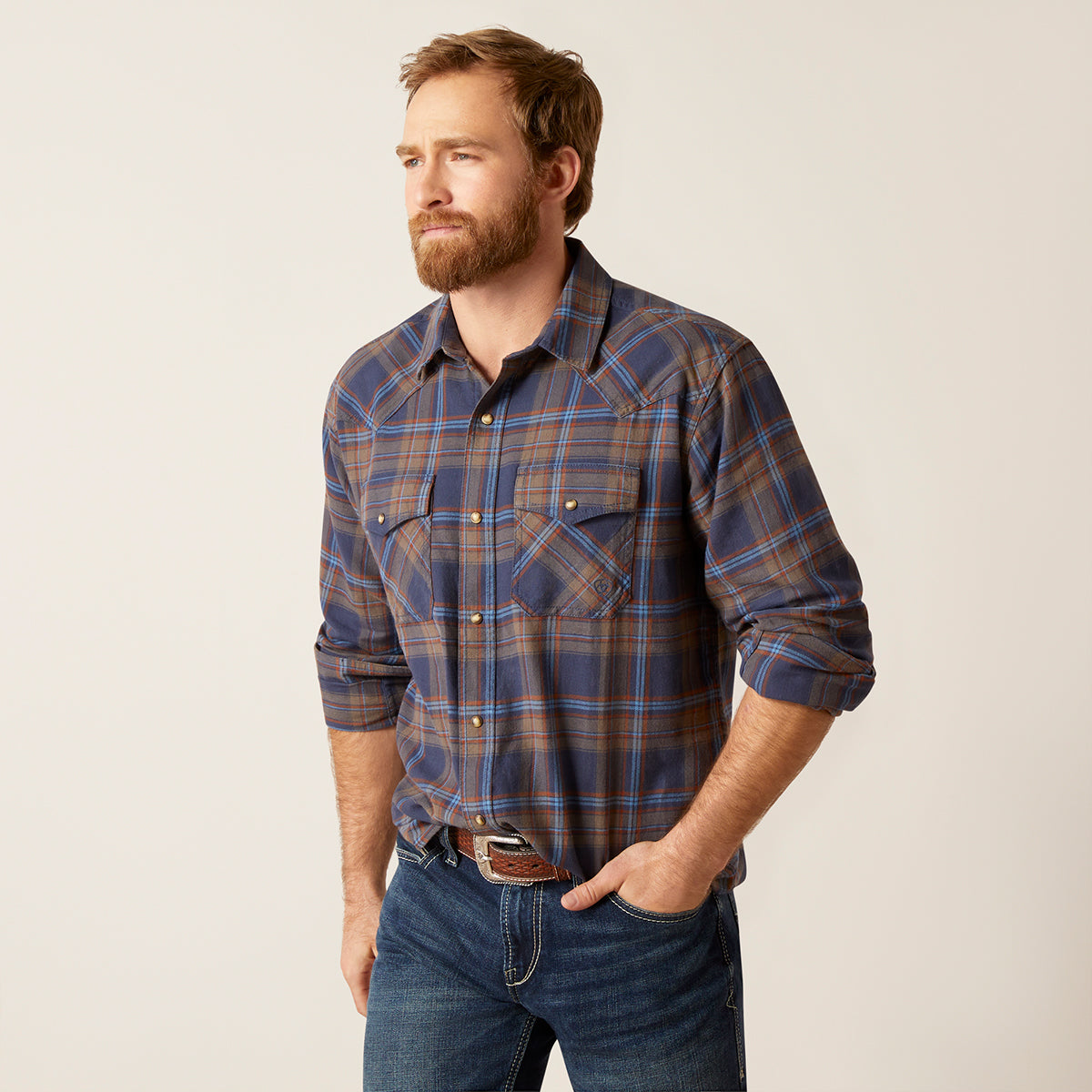 A man wearing a long-sleeve western-style plaid button-down Ariat shirt in navy, brown, and orange, paired with blue jeans, standing against a light beige studio background.