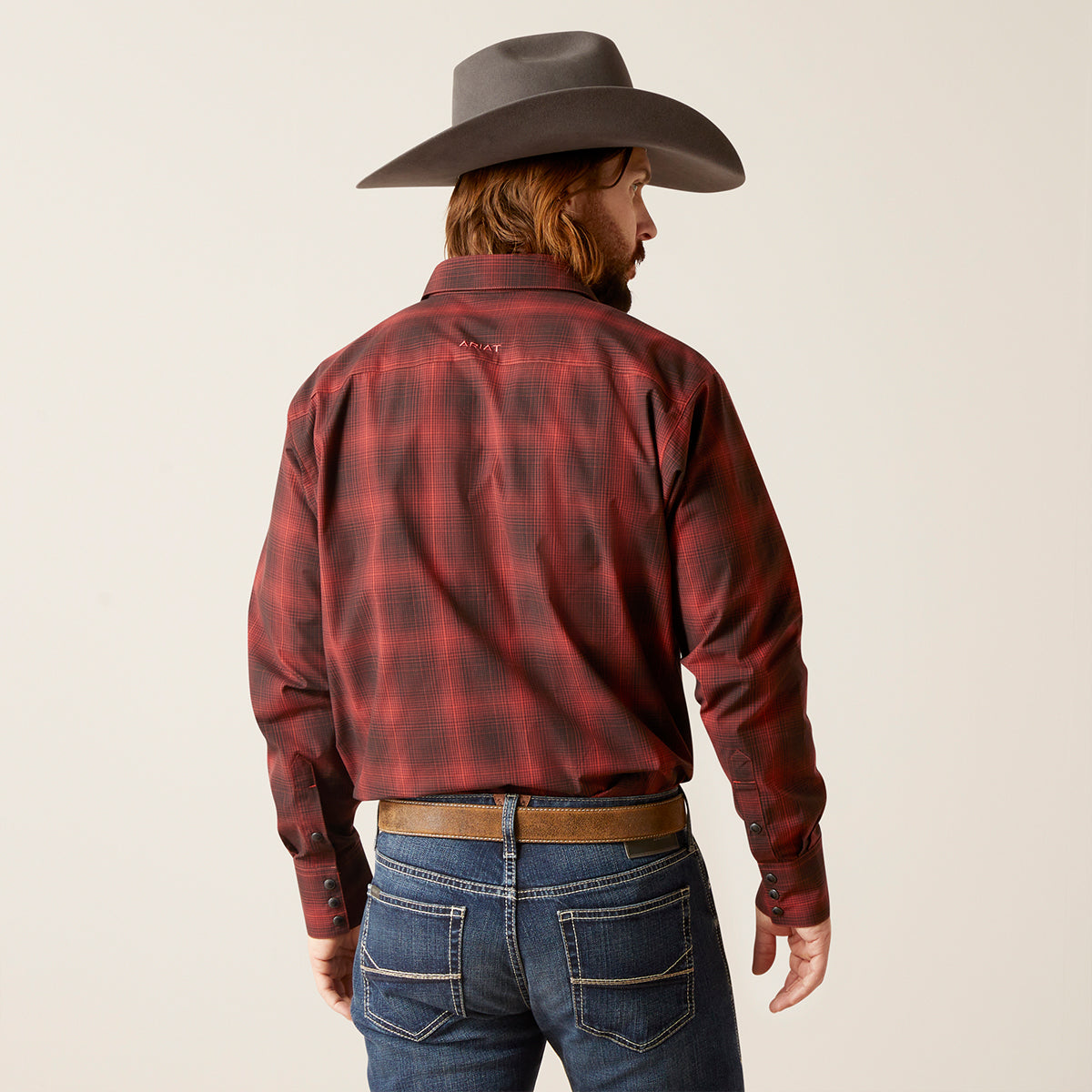 A man wearing a red and black plaid western Ariat shirt, blue jeans, a brown belt, and a dark brown cowboy hat, shown from the back against a light beige studio background.