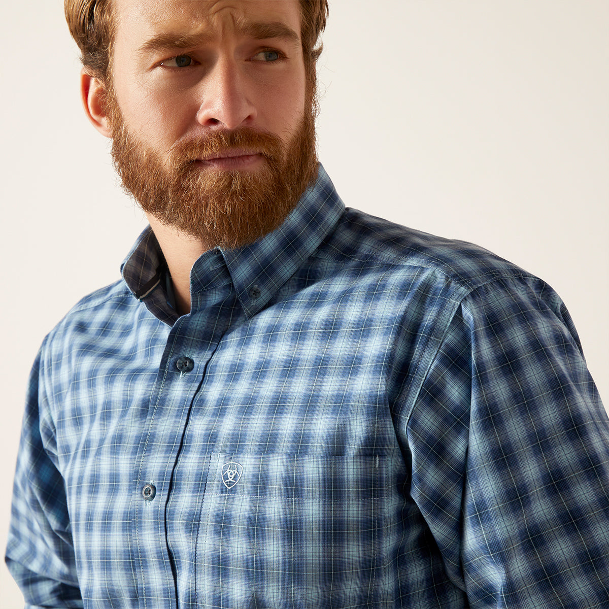 A close-up of a man wearing a blue and white plaid button-down shirt. The shirt features a classic checkered pattern in shades of blue and white, with a small embroidered logo on the left chest pocket.