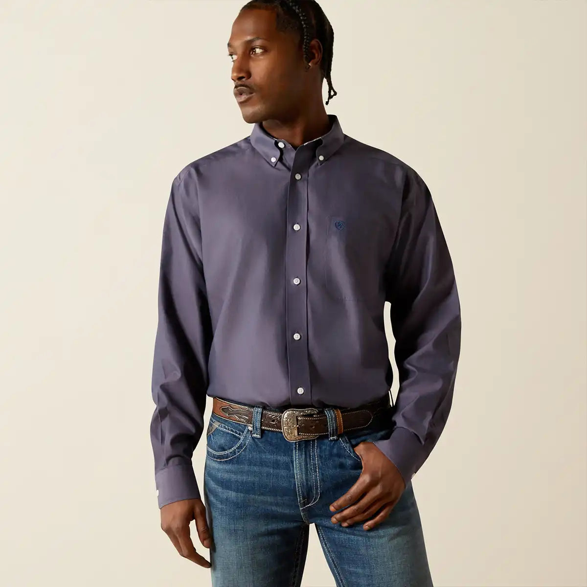 A man wearing a long-sleeve button-down Ariat shirt in a muted purple shade, paired with blue jeans and a brown belt, standing against a plain beige background.