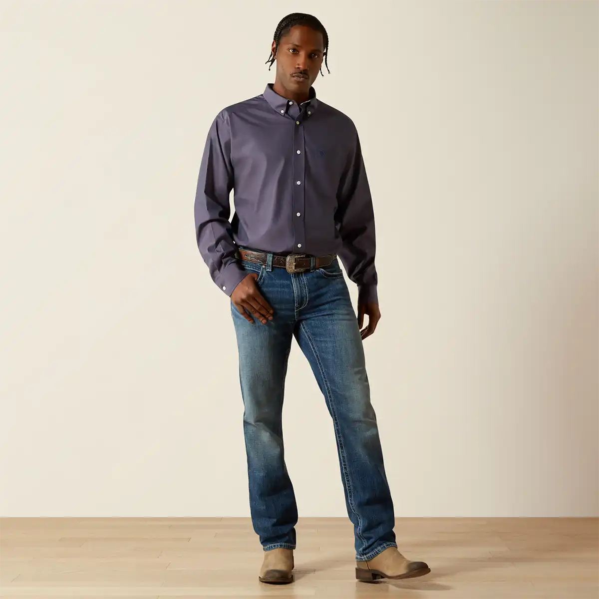 A man modeling a dark purple long-sleeve button-down Ariat shirt with blue jeans and brown boots, standing against a plain beige studio background.