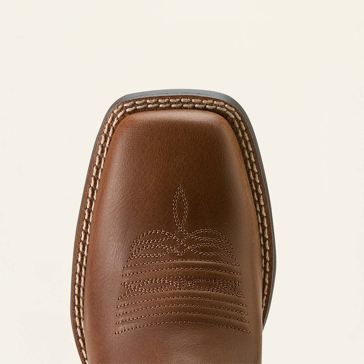 Close-up of the Ariat square toe of a brown leather cowboy boot with decorative stitching and a western-style pull tab, shown on a light cream background.