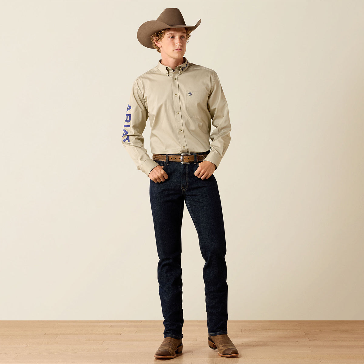 A man wearing a light beige western long-sleeve button-up Ariat shirt with blue vertical lettering on the sleeve, dark blue jeans, a brown belt, brown boots, and a brown cowboy hat, standing against a plain beige studio background.