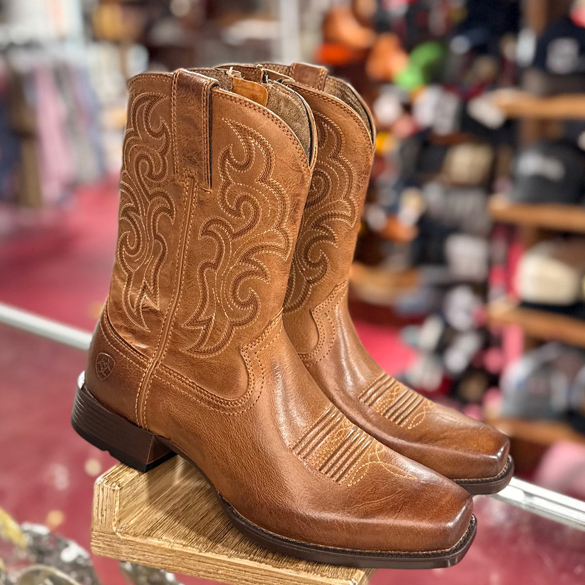 Pair of Ariat men's brown square toe cowboy boots on a wooden block in a store setting.