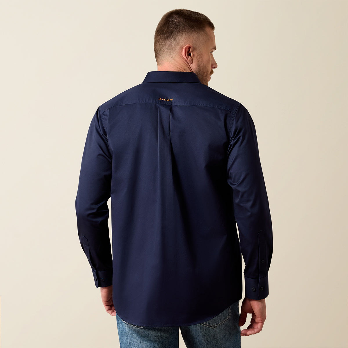 A man viewed from the back wearing a navy blue long-sleeve button-up shirt and blue jeans, standing against a plain beige background.