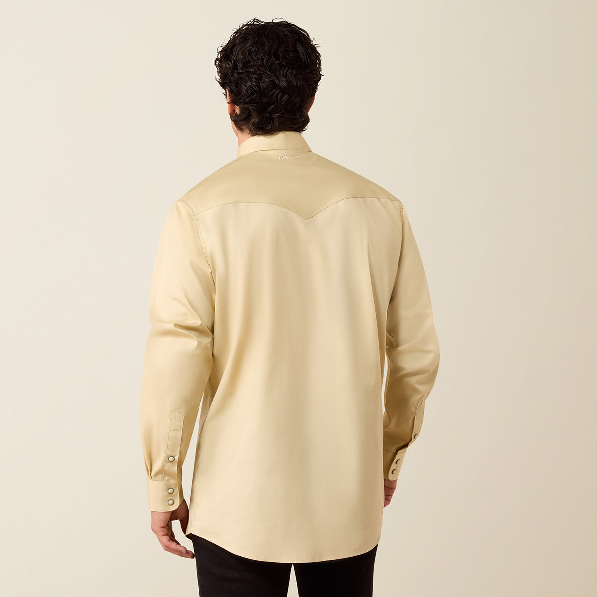 A man viewed from behind wearing a light beige long-sleeve western-style shirt with a curved yoke and black pants, set against a plain light beige studio background.