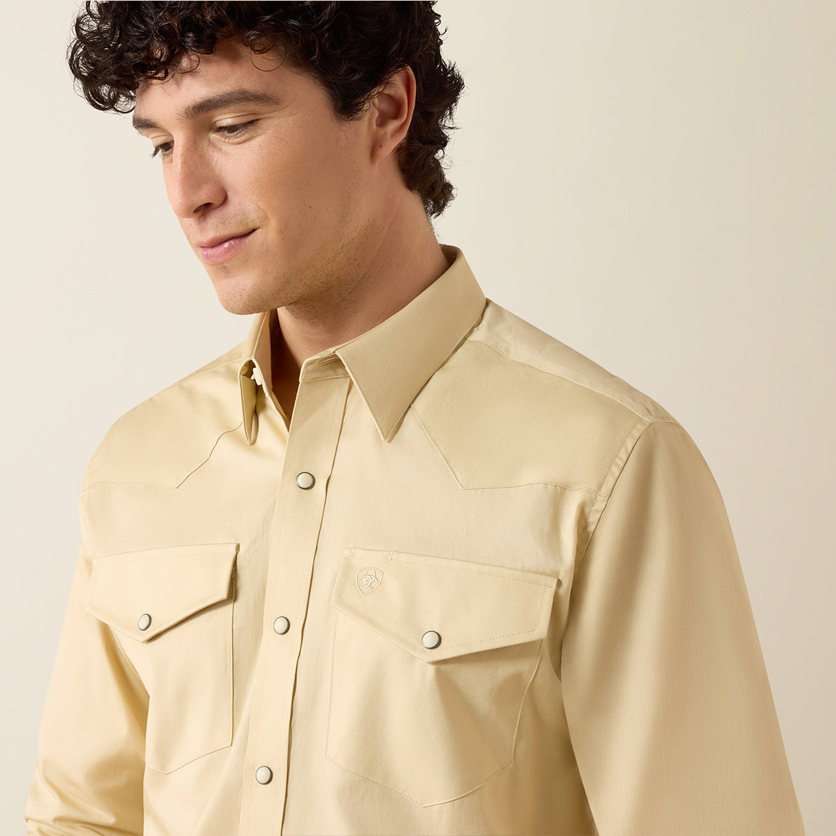 A man wearing a light beige western-style button-down shirt with two chest pockets, photographed against a plain light beige background.