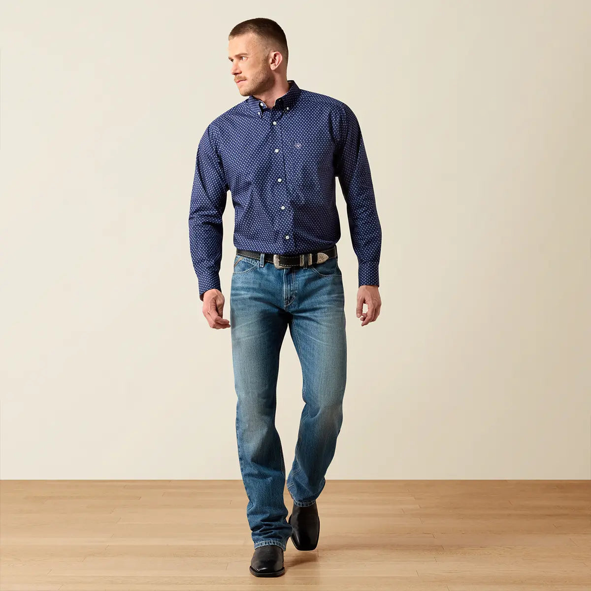Full-body studio lifestyle view of the male model wearing the Ariat navy geo print shirt, blue jeans, black Western belt, and black boots.