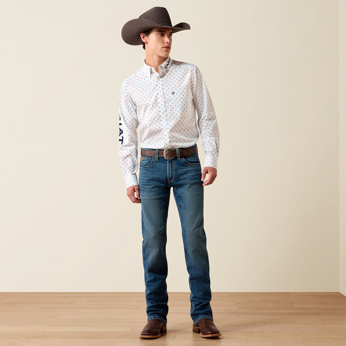 A man wearing Ariat white long-sleeve western shirt with a small repeating pattern, blue jeans, a brown cowboy hat, and brown boots, standing against a plain beige wall and light wood floor.