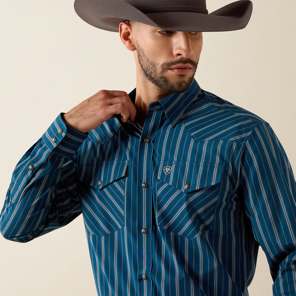 A man wearing a dark brown cowboy hat and a blue western-style button-down shirt with thin white vertical stripes. He is adjusting the collar of his shirt against a plain beige background.