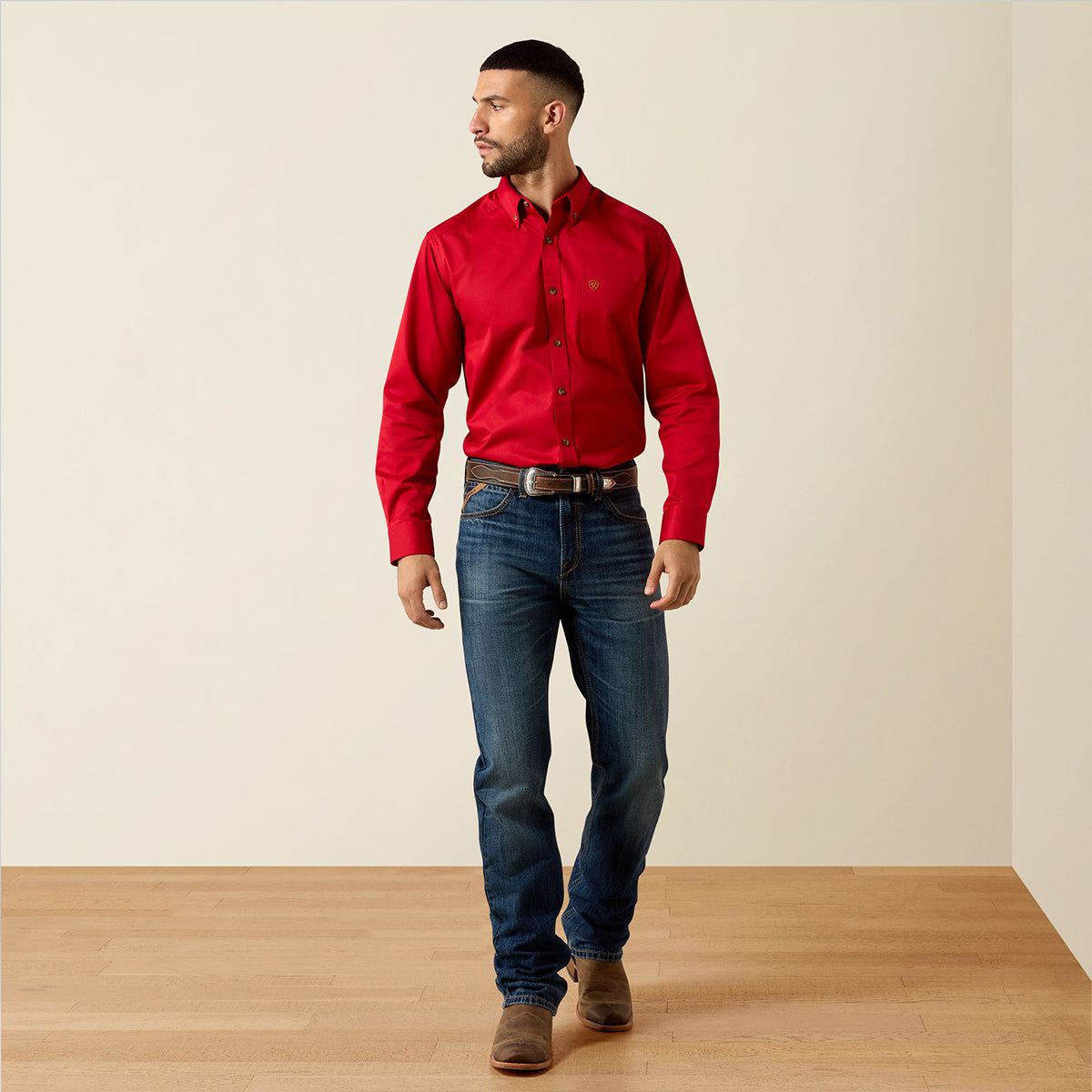 A man wearing a Ariat bright red long-sleeve button-down shirt, dark blue jeans, and brown boots, standing in a studio with a beige wall and light wood floor.