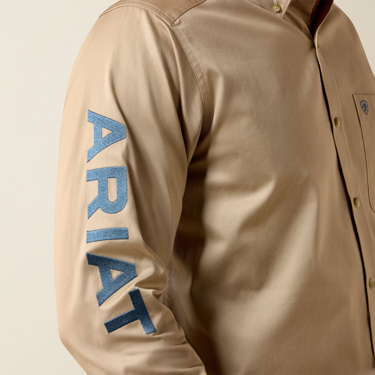 Close-up of a beige long-sleeve button-up shirt worn by a model, featuring a large blue 'ARIAT' logo printed vertically on the right sleeve.
