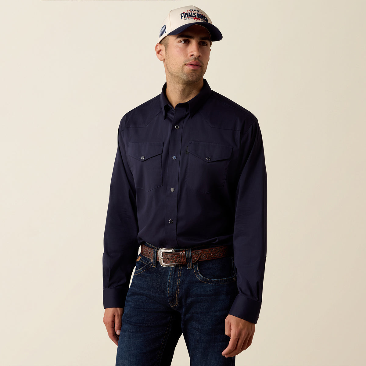A man wearing Ariat dark navy long-sleeve western-style button-down shirt, dark blue jeans, a brown patterned belt, and a white baseball cap with a 'Pima' logo, standing against a plain beige background.
