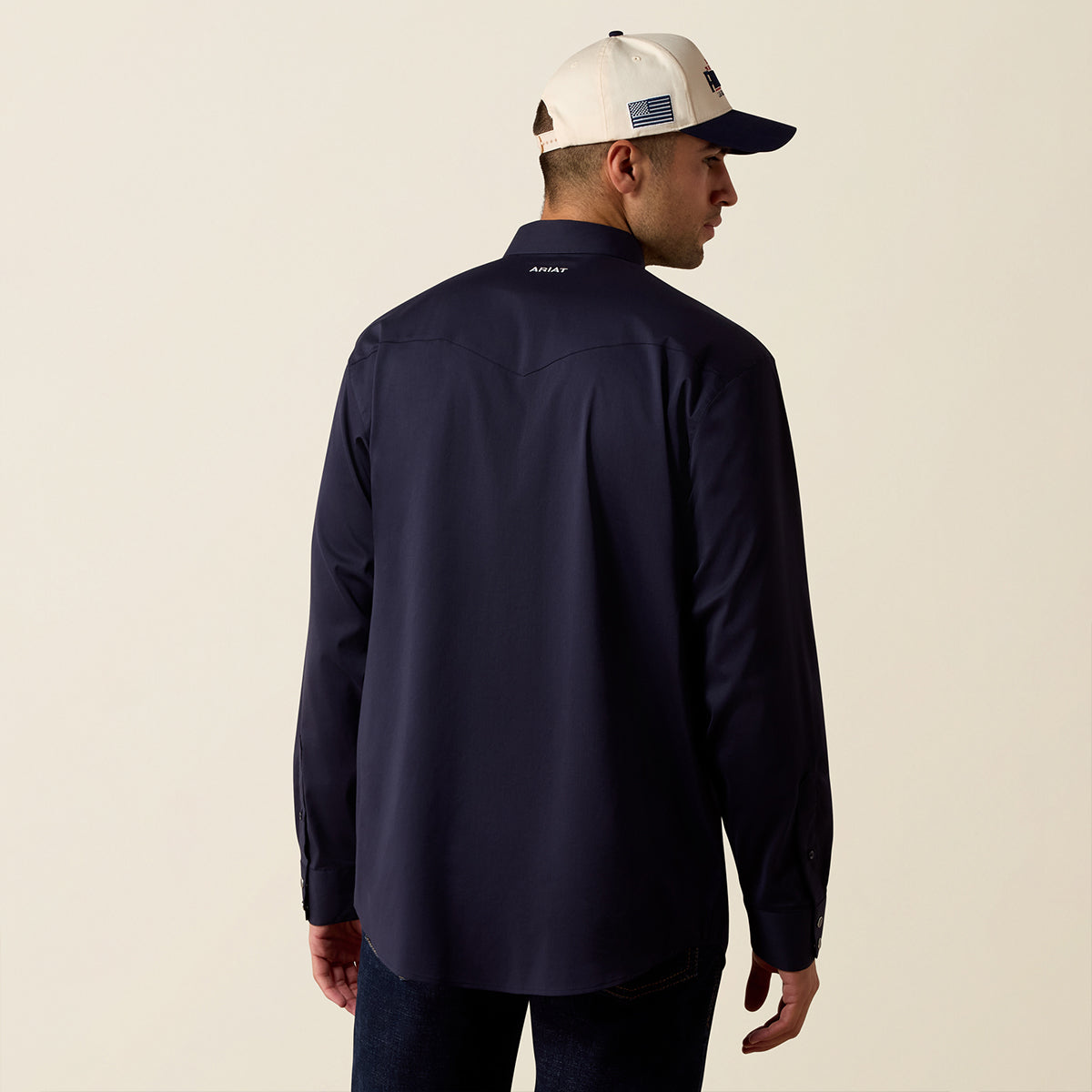 A man wearing a navy blue long-sleeve performance shirt and a cream and navy baseball cap, shown from the back against a light beige studio background.