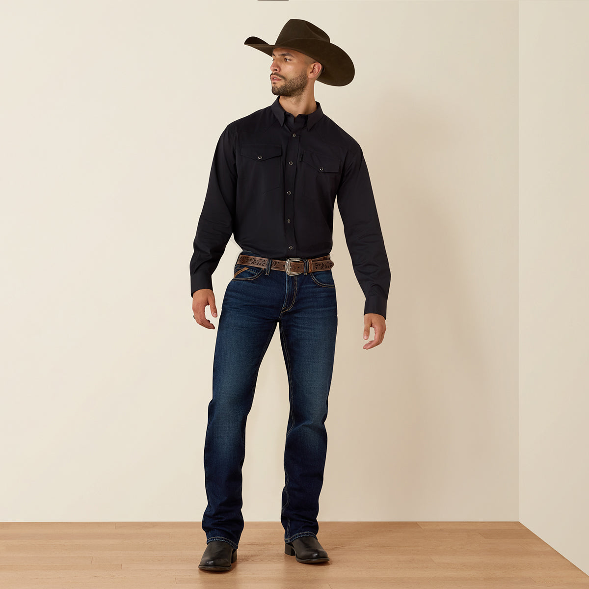 A man wearing an Ariat black long-sleeve button-down shirt, dark blue jeans, a brown western belt, and a brown cowboy hat, standing against a plain beige wall and light wood floor.