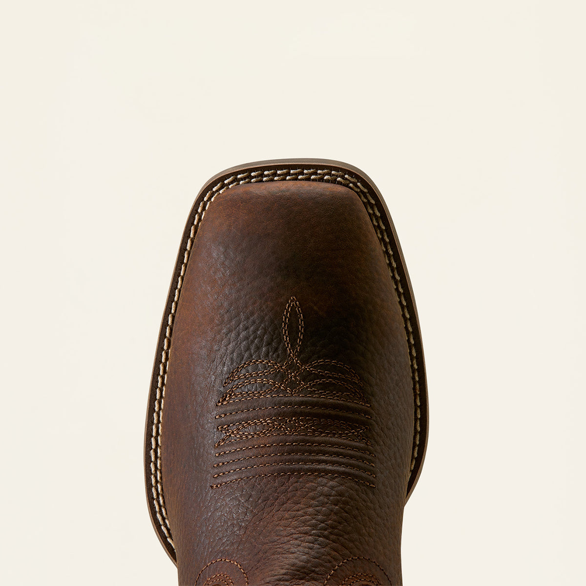 Close-up of the Ariat square toe and vamp of a dark brown leather cowboy boot with decorative stitching on a light cream background.