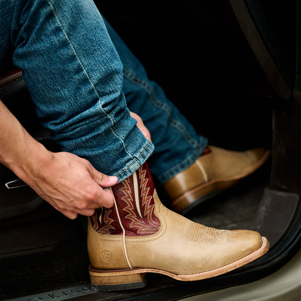 A tan leather Ariat cowboy boot with red western embroidery being put on a person's foot inside a car, with blue jeans visible.