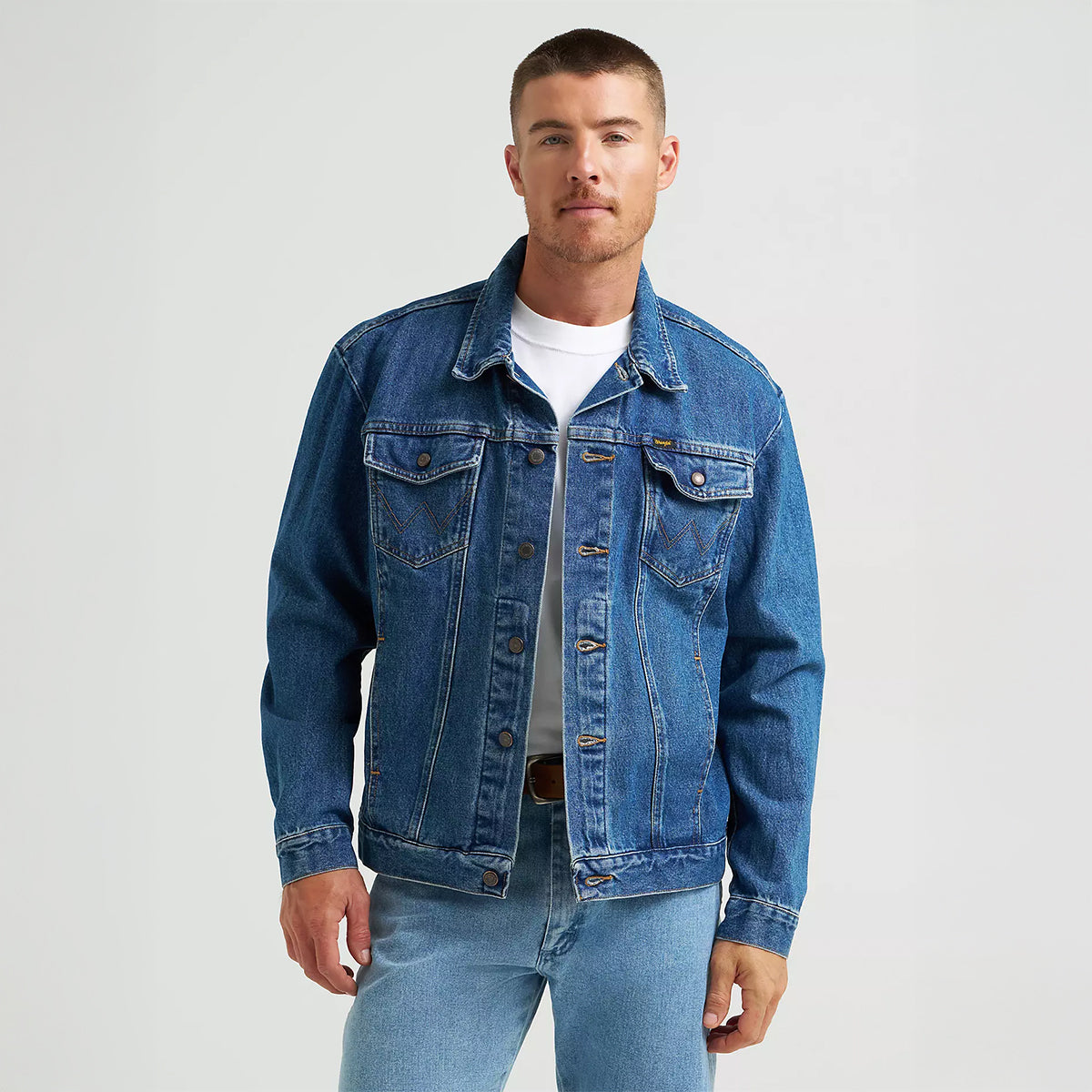 Front view of a model wearing the Wrangler Men's Cowboy Cut unlined denim jacket in medium stonewash blue layered over a white t-shirt.